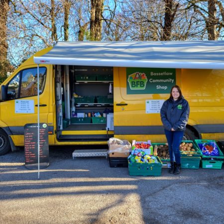 A photo of the mobile Bassetlaw Community Shop which is a bright yellow van. In the photo crates of food are shown outside the van whilst a woman is standing and smiling in front of the van.