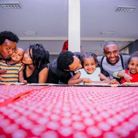 A group pf smiling adults and children gathered around a table with a red and white checkered tablecloth on it