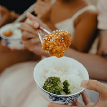 A photo of a piece of sticky meat coated in sauce being held in chopsticks above a small bowl of white rice
